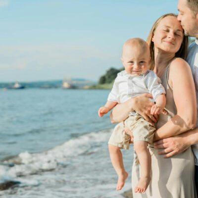 A joyful family moment captured by the sea, with a couple holding their smiling baby on a sunny day.