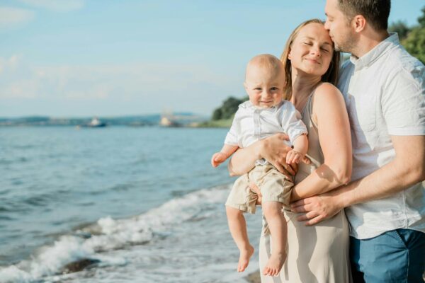 A joyful family moment captured by the sea, with a couple holding their smiling baby on a sunny day.