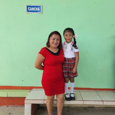 A woman and her smiling daughter in school uniform pose for a picture outdoors near a school building.