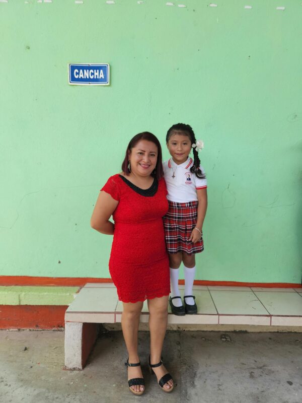 A woman and her smiling daughter in school uniform pose for a picture outdoors near a school building.