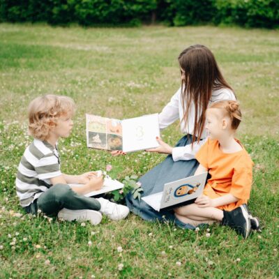 Two children and a woman enjoy a sunny outdoor reading session on the grass, enhancing their learning experience.