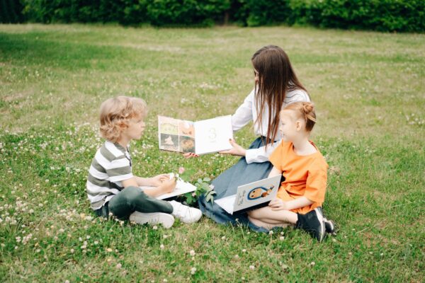 Two children and a woman enjoy a sunny outdoor reading session on the grass, enhancing their learning experience.